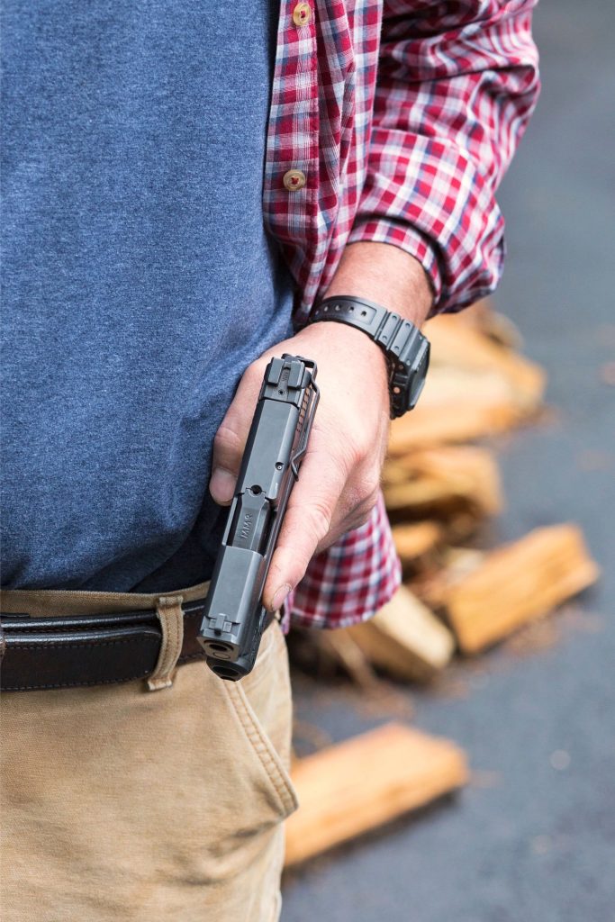 A person wearing a plaid shirt and tan pants drawing a handgun with his non-dominant hand using a Clipdraw gun clip for concealed carry.